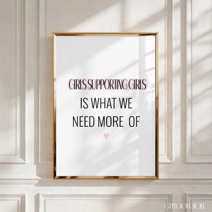 A framed typographic wall art print leaning against a white panelled wall. The print has a gold frame and reads, 'GIRLS SUPPORTING GIRLS IS WHAT WE NEED MORE OF' with a small pink heart below.