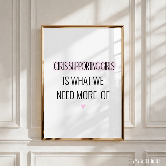 A framed typographic wall art print leaning against a white panelled wall. The print has a gold frame and reads, 'GIRLS SUPPORTING GIRLS IS WHAT WE NEED MORE OF' with a small pink heart below.