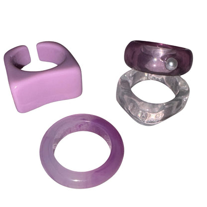 High-angle view of a four-piece purple and clear acrylic ring stack spread out on a pink journal, featuring a chunky lilac signet ring, a marbled purple band, a wavy clear ring, and a translucent purple dome ring with a faux pearl.