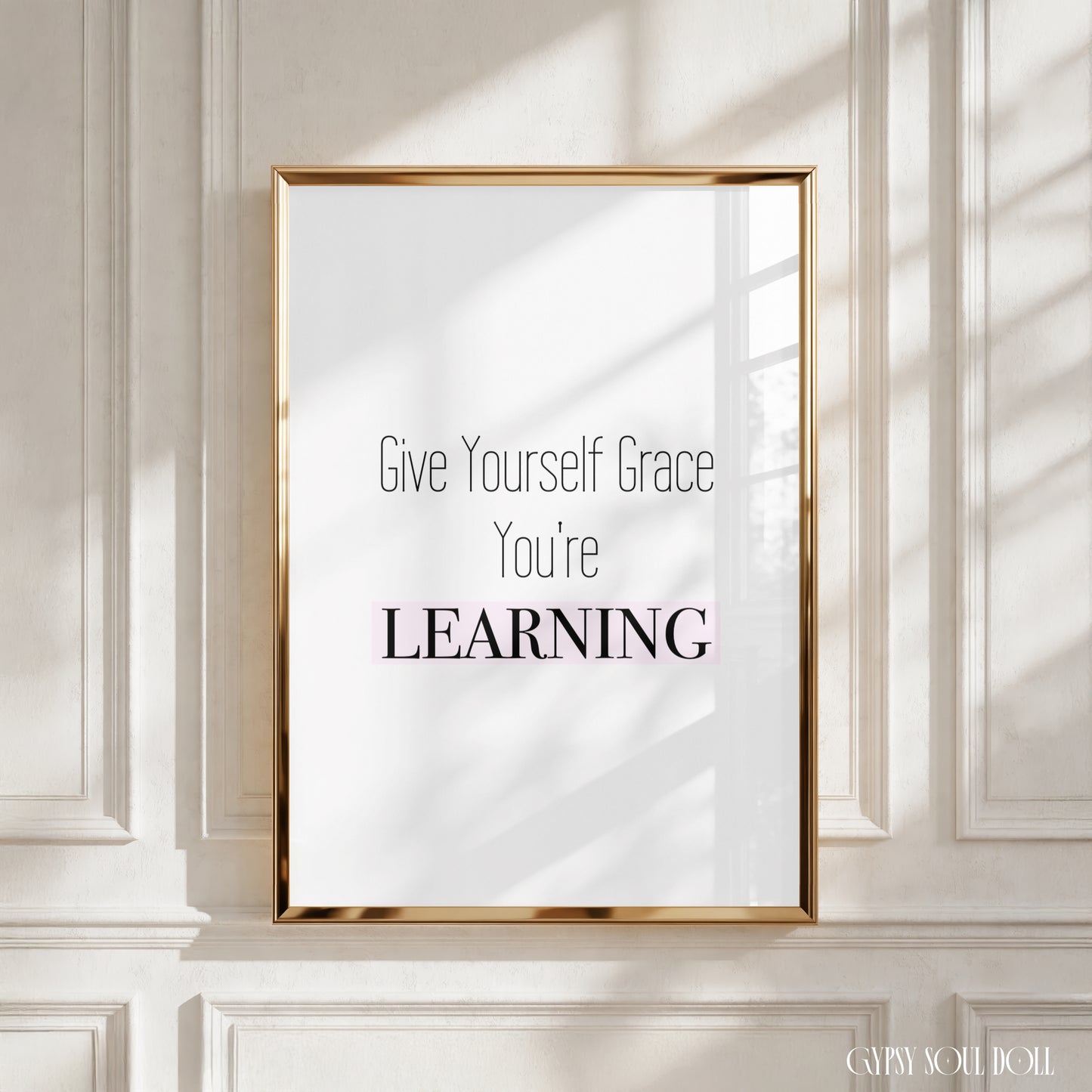 A framed typographic wall art print with the text 'Give Yourself Grace You're LEARNING' in black and pink lettering on a white background, displayed against a cream paneled wall with soft window-shadow light.