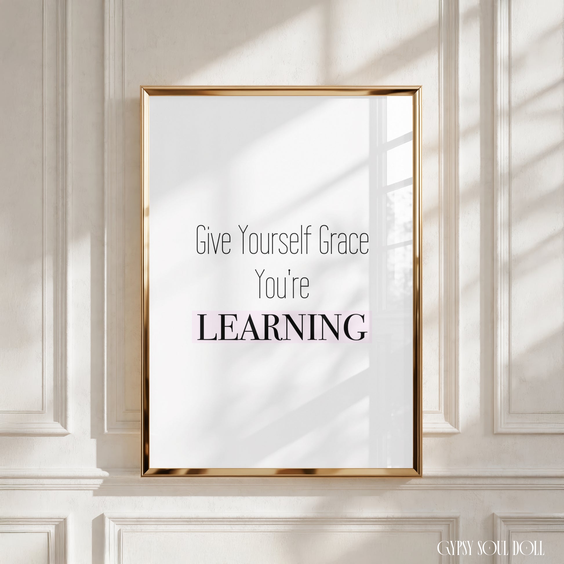A framed typographic wall art print with the text 'Give Yourself Grace You're LEARNING' in black and pink lettering on a white background, displayed against a cream paneled wall with soft window-shadow light.