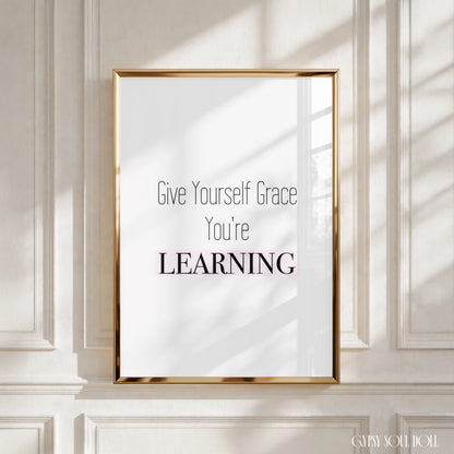A framed typographic wall art print with the text 'Give Yourself Grace You're LEARNING' in black and pink lettering on a white background, displayed against a cream paneled wall with soft window-shadow light.