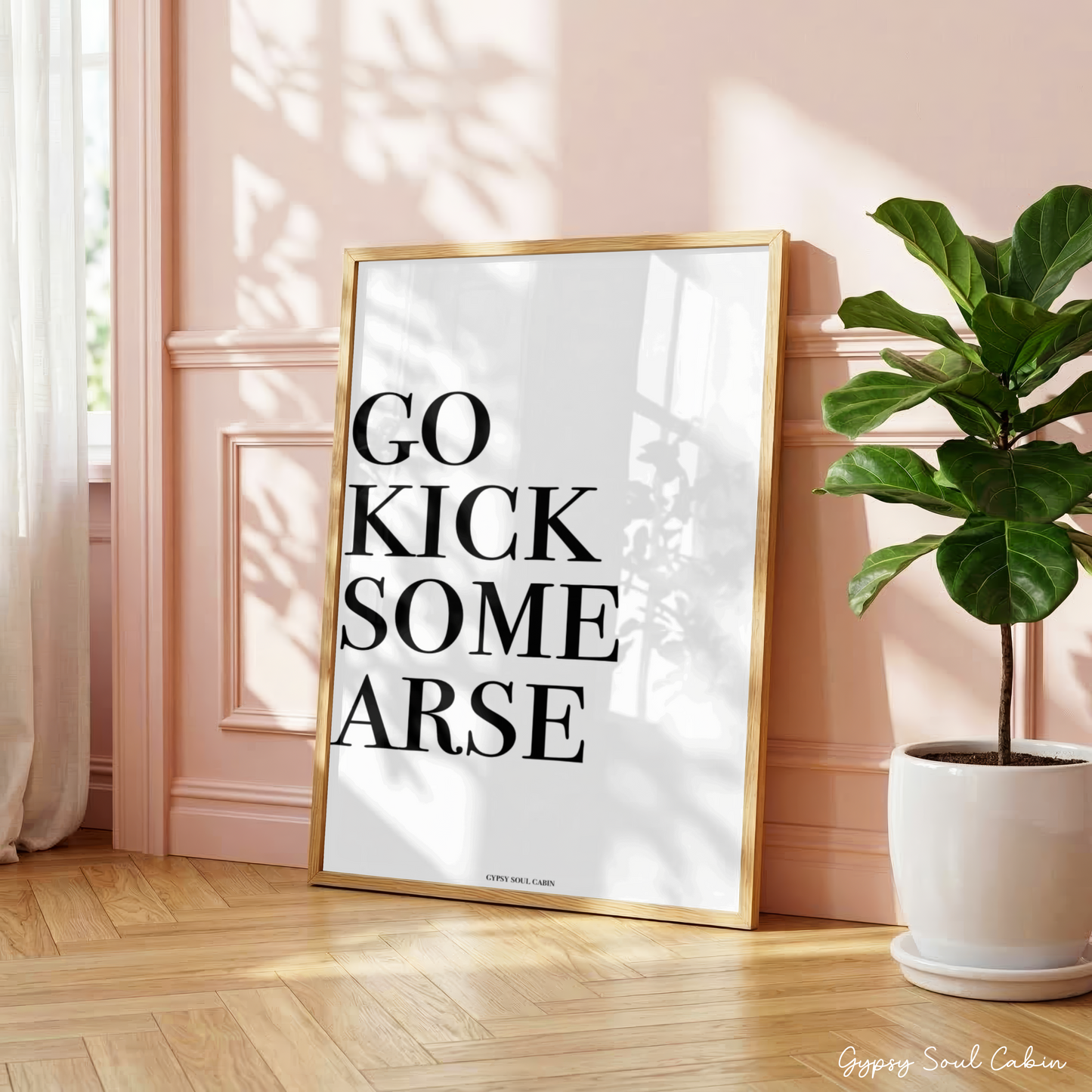 Minimalist black and white manifestation affirmation typography print reading Go Kick Some Arse in a large oak frame leaning against a panelled pink wall.