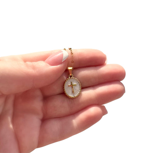 Close-up view of a hand with a classic white French tip manicure holding a gold oval cross medallion pendant necklace with a shimmering vanilla enamel background.