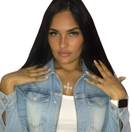 A model with long dark hair wearing a light blue denim jacket over a white top, featuring a silver cross pendant necklace. She is posed against a solid white background.