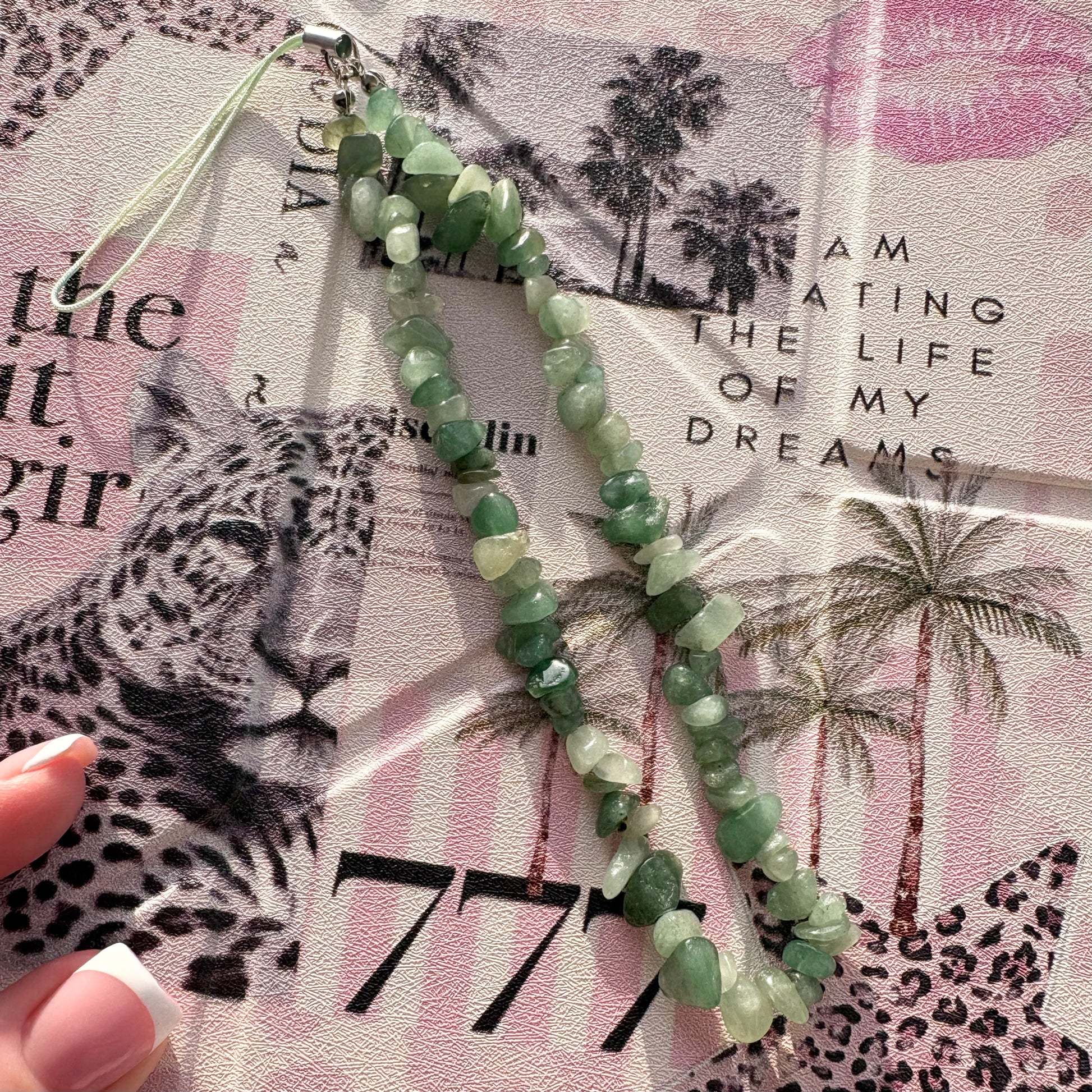 A beaded phone strap made of green faceted gemstone-like beads with a silver metal clasp and loop, laid over a pink leopard-print paper background. A hand with a French manicure is partially visible at the lower left.