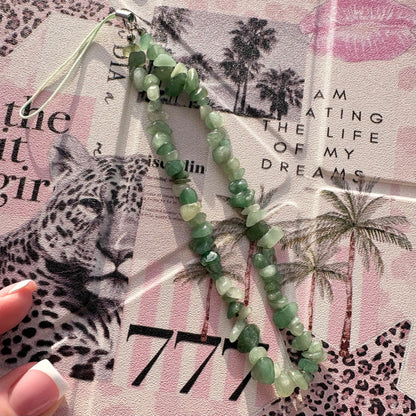 A beaded phone strap made of green faceted gemstone-like beads with a silver metal clasp and loop, laid over a pink leopard-print paper background. A hand with a French manicure is partially visible at the lower left.