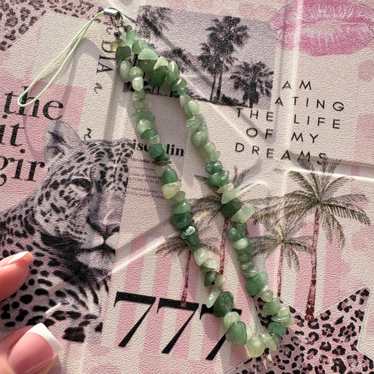 A beaded phone strap made of green faceted gemstone-like beads with a silver metal clasp and loop, laid over a pink leopard-print paper background. A hand with a French manicure is partially visible at the lower left.