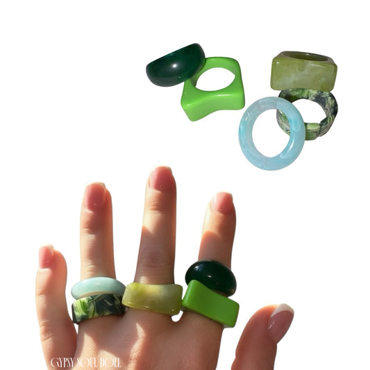 A hand with a classic French manicure modelling a five-piece stack of chunky Y2K style acrylic rings in shades of bright lime green, dark emerald, marbled olive, and icy translucent blue, with a digital cutout of the rings floating above against a clean white background stamped with the Gypsy Soul Doll watermark