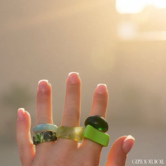 A close-up of a hand wearing four colourful rings under warm golden sunlight. The rings include a light green stone, a mixed green marbled band, a translucent green stone, and a bright lime green stone.