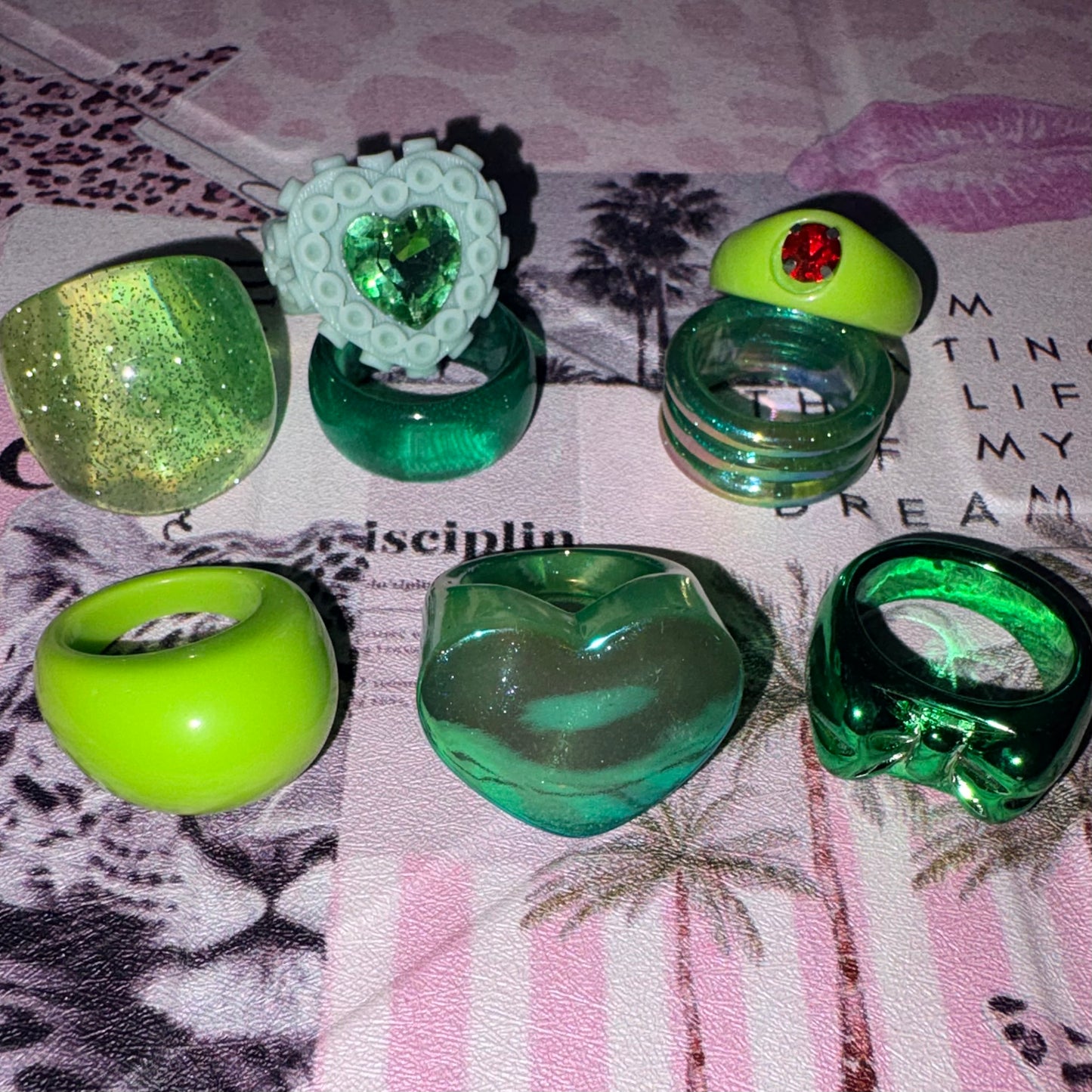 Eight chunky green retro rings, including a heart and a bow, arranged on a pink-and-white patterned fabric background.