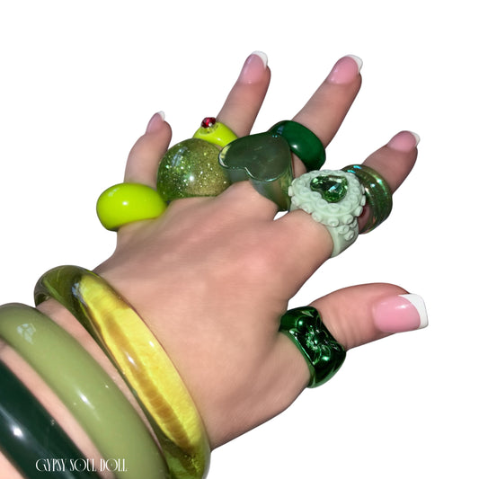 A hand wearing multiple chunky green rings and matching green bangles, photographed against a plain white background. The rings include glossy green, translucent green, textured white with green accents, and one with a small red detail.