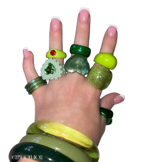 A hand with manicured nails wearing multiple chunky green rings and matching green bangles, photographed against a plain white background. The rings feature various green textures and stones, including a textured white ring with a green gemstone and a green glitter ring.