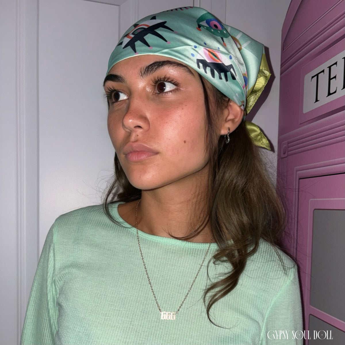 A model wearing a green evil eye silk boho headscarf paired with a matching mint green ribbed long-sleeve top and an Angel number 666 gold necklace.