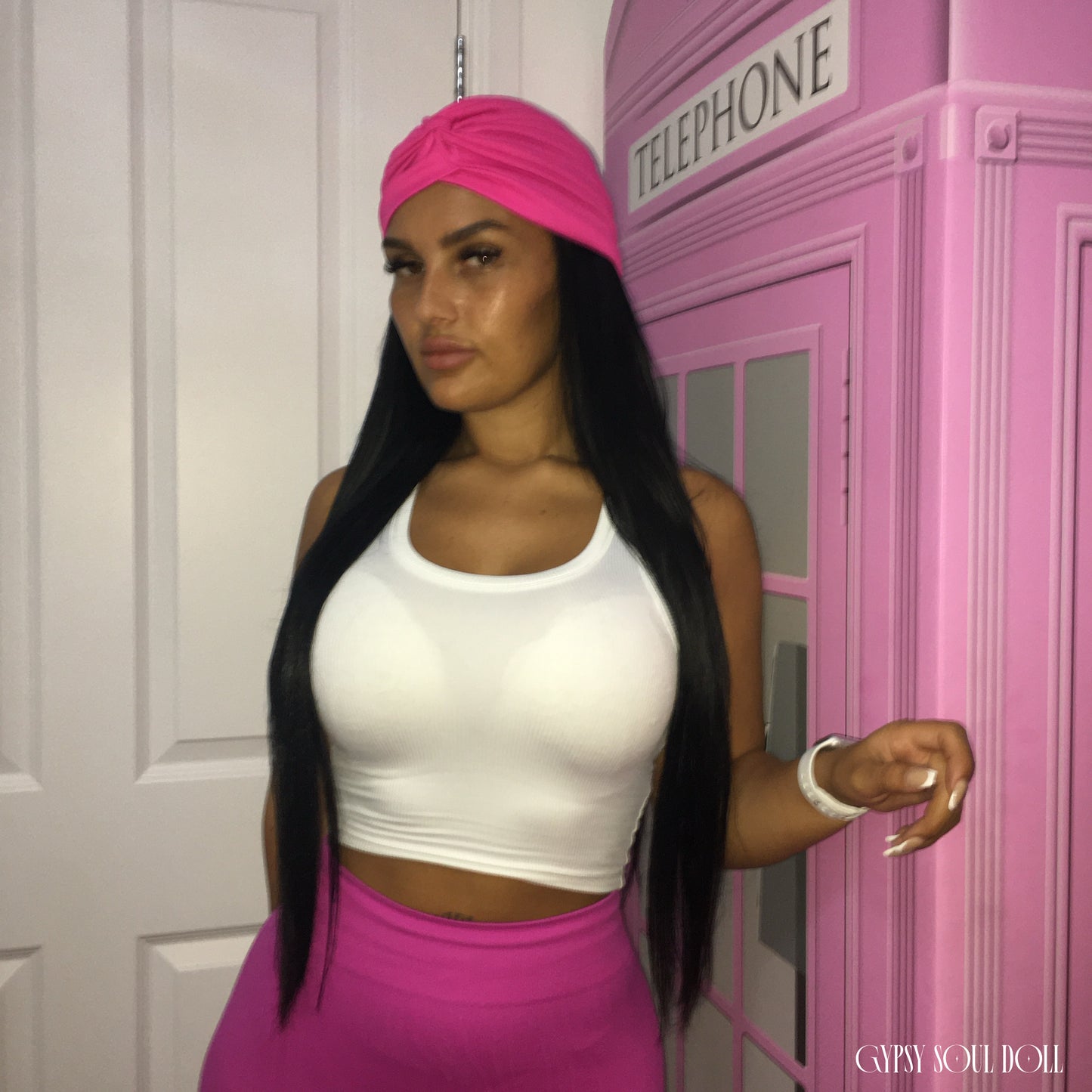 A woman posing in a white crop top and bright pink skirt, wearing a matching pink headwrap. She is standing next to a pink telephone booth prop.