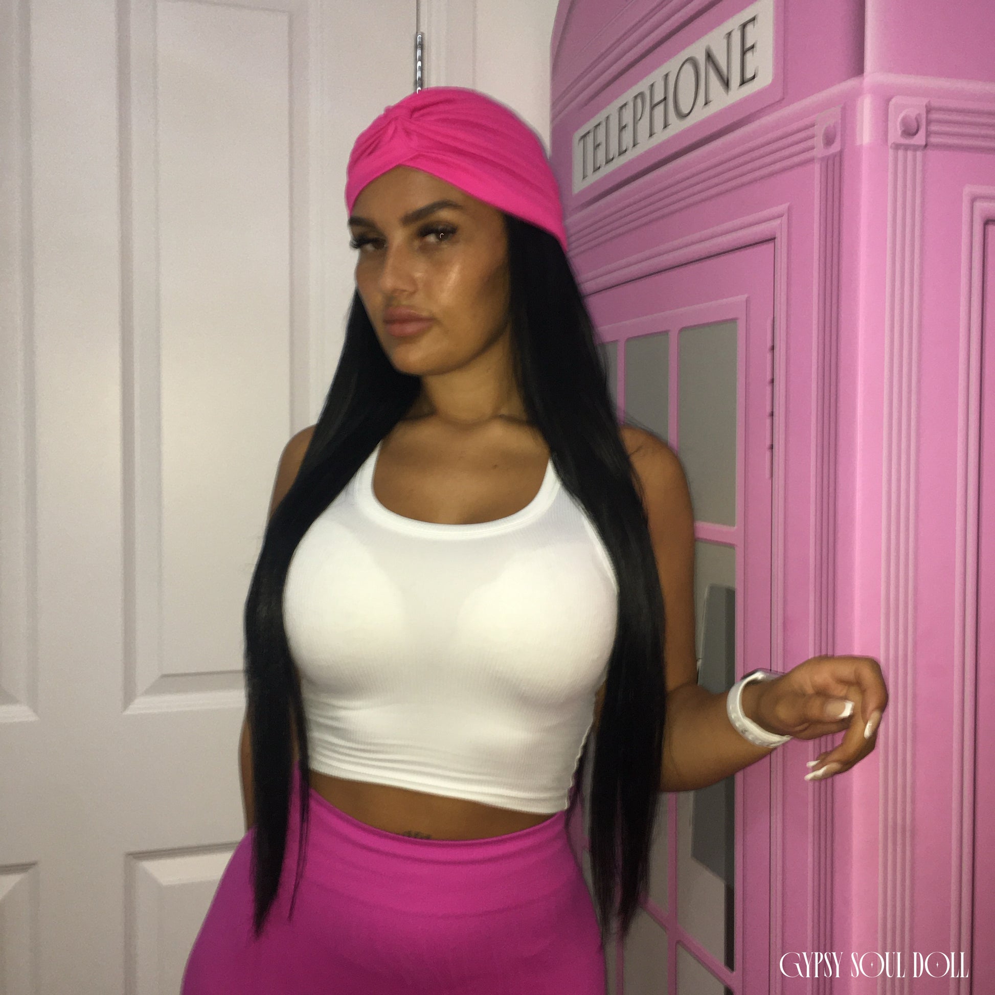 A woman posing in a white crop top and bright pink skirt, wearing a matching pink headwrap. She is standing next to a pink telephone booth prop.