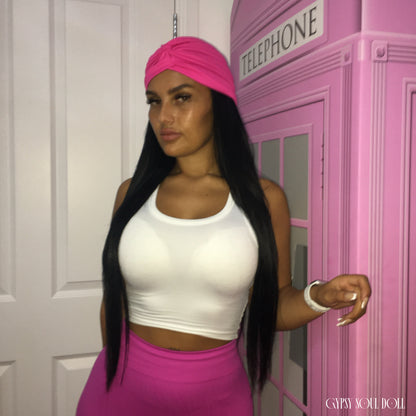 A woman posing in a white crop top and bright pink skirt, wearing a matching pink headwrap. She is standing next to a pink telephone booth prop.
