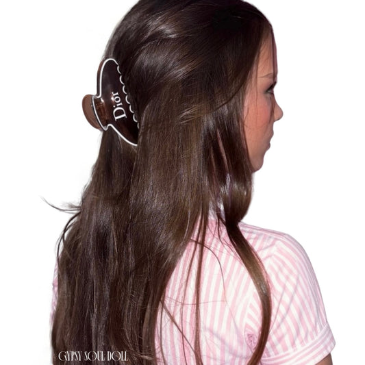 A model is shown from the back and side, wearing a large brown hair claw clip with a white scalloped border and the word "Dior" printed on it. She has long, straight brown hair and is wearing a pink and white striped shirt against a plain white background.