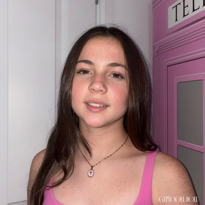 Front view of a model with long brown hair wearing a pink top next to a pink telephone box display, showcasing the Gypsy Soul Doll dainty gold link necklace featuring an oval cross medallion pendant with a pink inlay.