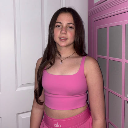 Front view of a model with long brown hair wearing a pink top next to a pink telephone box display, showcasing the Gypsy Soul Doll dainty gold link necklace featuring an oval cross medallion pendant with a pink inlay.