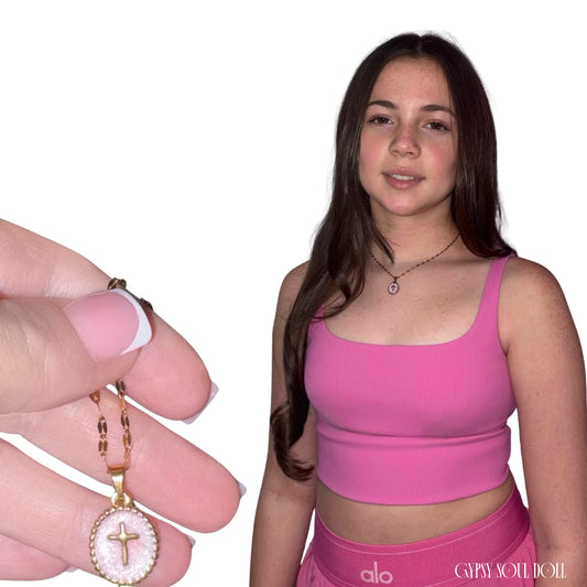 Front view of a model with long brown hair wearing a pink top next to a pink telephone box display, showcasing the Gypsy Soul Doll dainty gold link necklace featuring an oval cross medallion pendant with a pink inlay.
