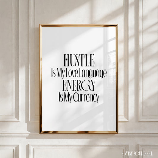 A framed typographic wall art print with the quote 'HUSTLE Is My Love Language ENERGY Is My Currency' in black serif text on a white background, set in a gold frame and hung on a cream panelled wall with soft window shadows by Gypsy Soul Doll.