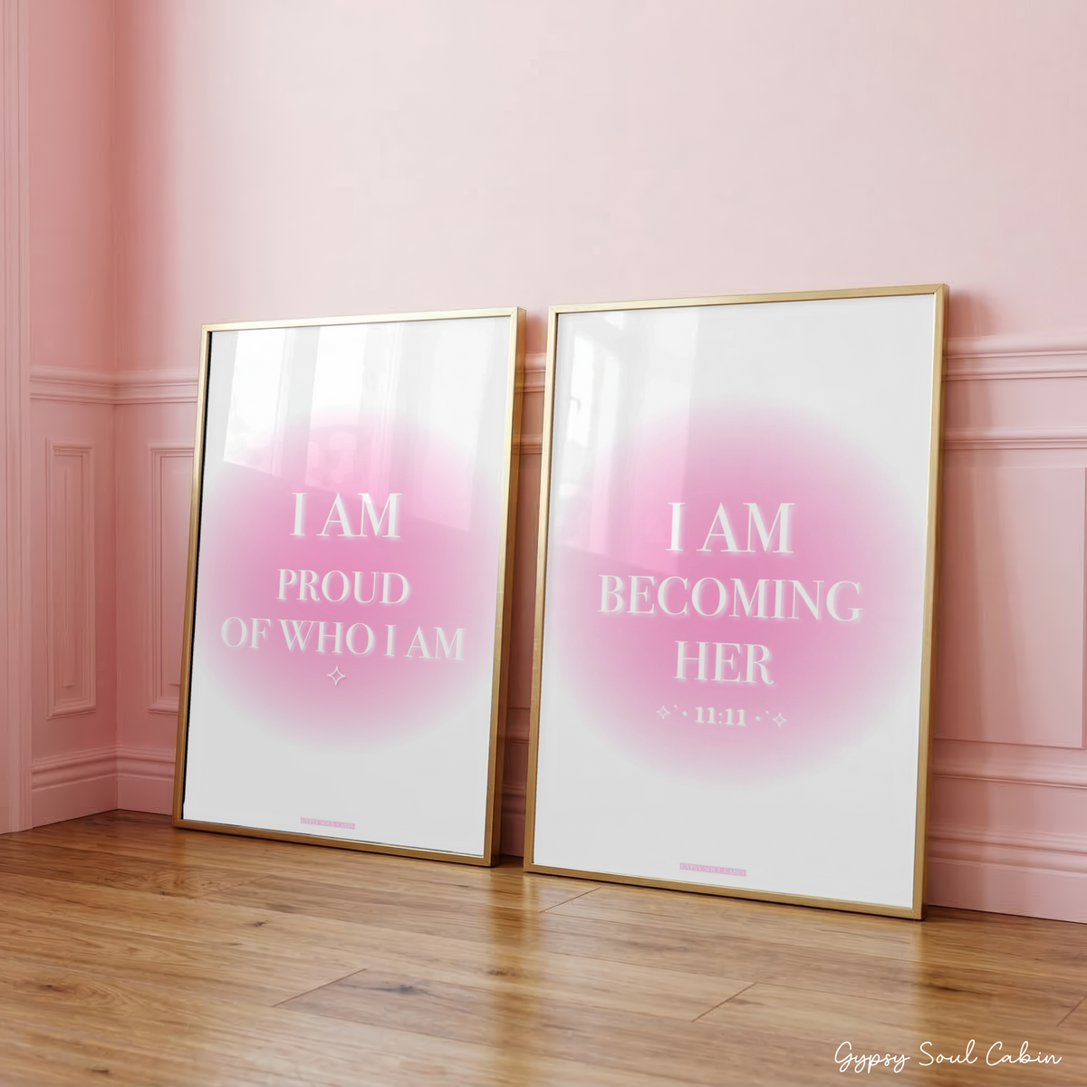 Two pink gradient aura typography prints reading "I Am Proud Of Who I Am" and "I Am Becoming Her 1111" in gold frames, leaning against a panelled pink wall.