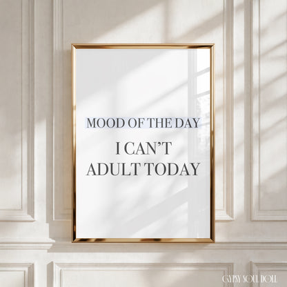 A framed typographic wall art print with the text 'MOOD OF THE DAY I CAN'T ADULT TODAY' in dark lettering on a white background, set in a thin gold frame and leaning against a white paneled wall with soft window shadows.