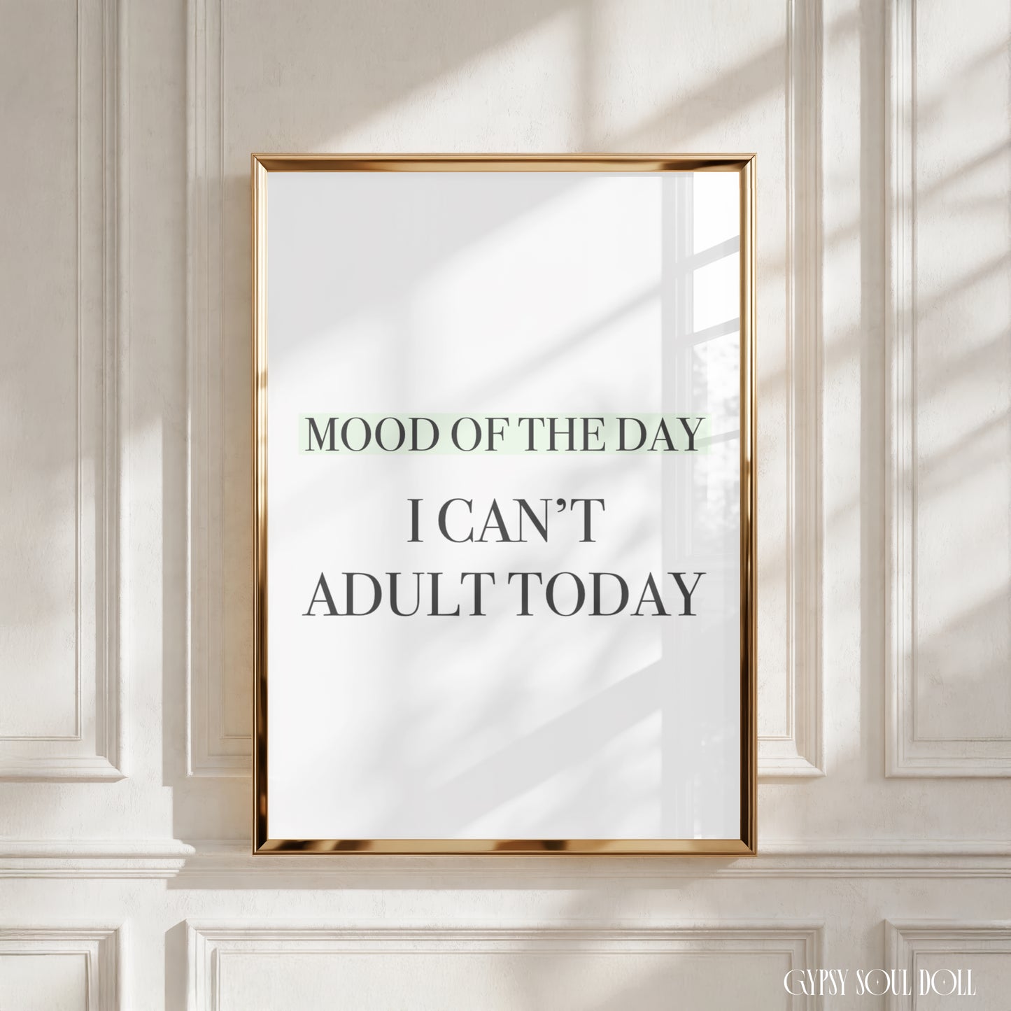 A framed typographic wall art print with the text 'MOOD OF THE DAY I CAN'T ADULT TODAY' hangs on a white paneled wall. The print has a white background with dark gray and muted green lettering, enclosed in a thin metallic gold frame.