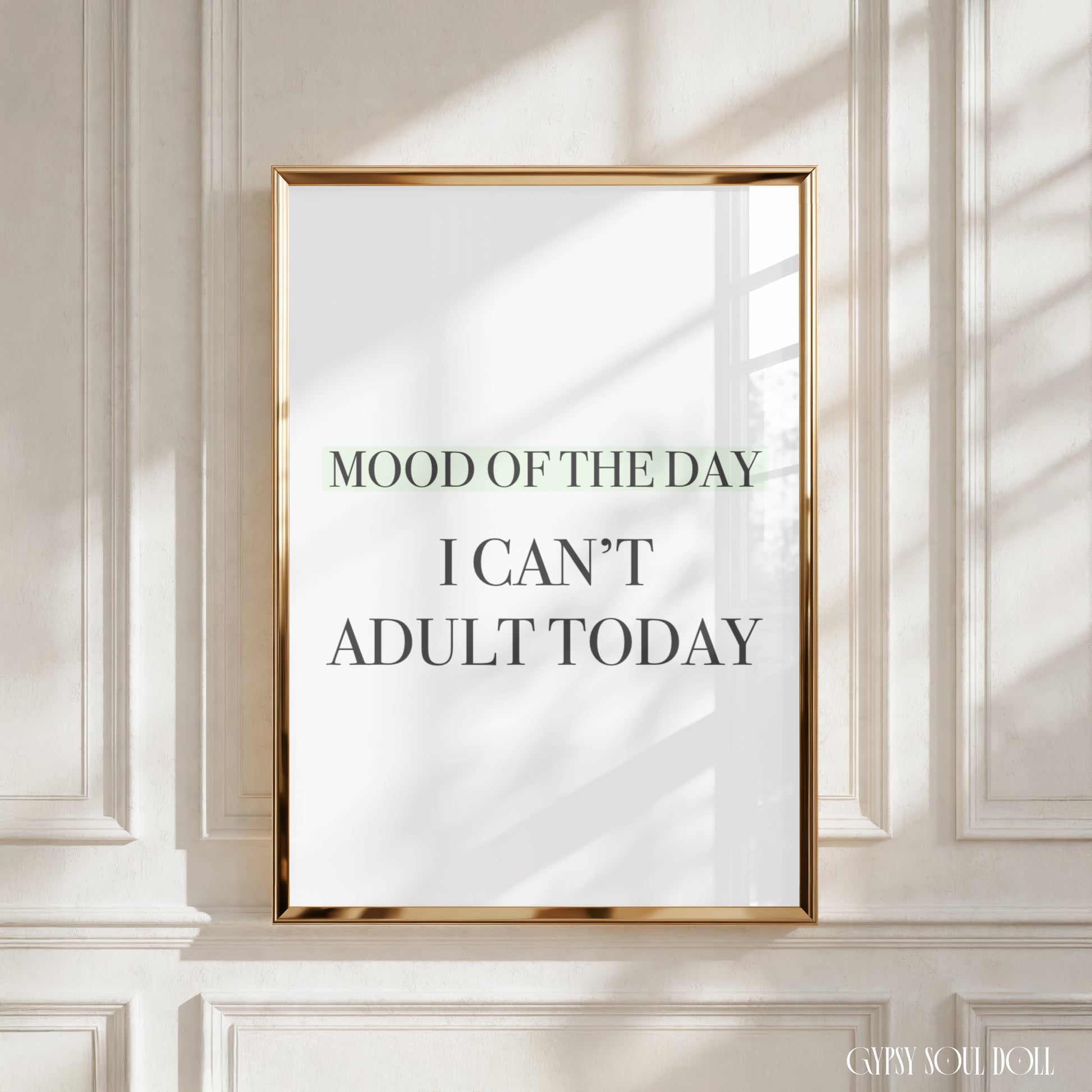 A framed typographic wall art print with the text 'MOOD OF THE DAY I CAN'T ADULT TODAY' hangs on a white paneled wall. The print has a white background with dark gray and muted green lettering, enclosed in a thin metallic gold frame.