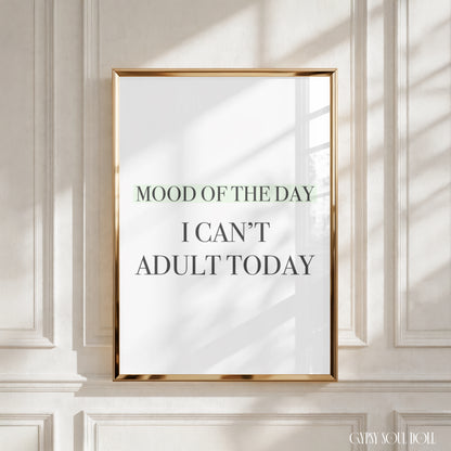 A framed typographic wall art print with the text 'MOOD OF THE DAY I CAN'T ADULT TODAY' hangs on a white paneled wall. The print has a white background with dark gray and muted green lettering, enclosed in a thin metallic gold frame.