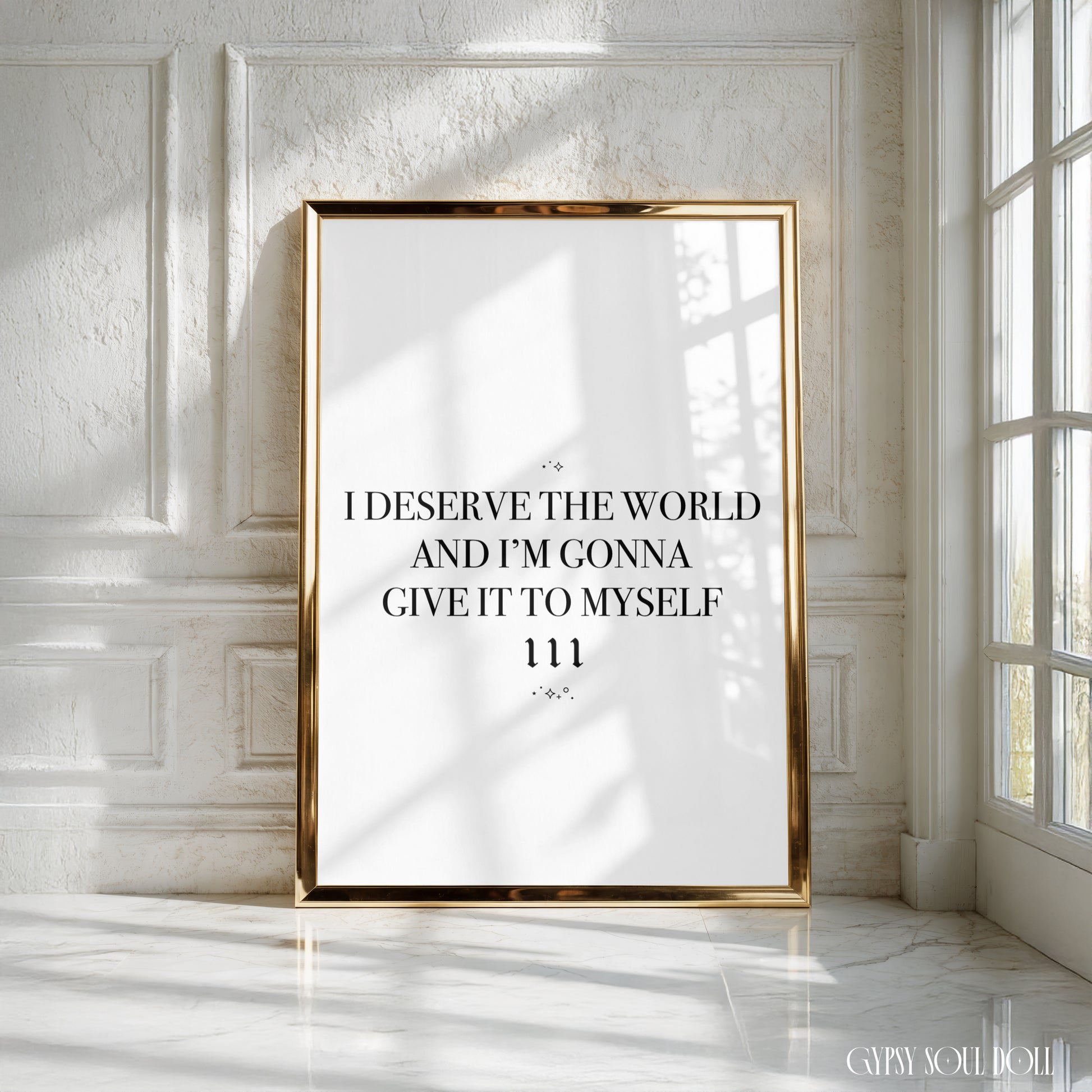 A vertical framed typographic art print with a gold frame leans against a white paneled wall in a sunlit room. The print has a white background with black serif text reading, 'I DESERVE THE WORLD AND I'M GONNA GIVE IT TO MYSELF 111.