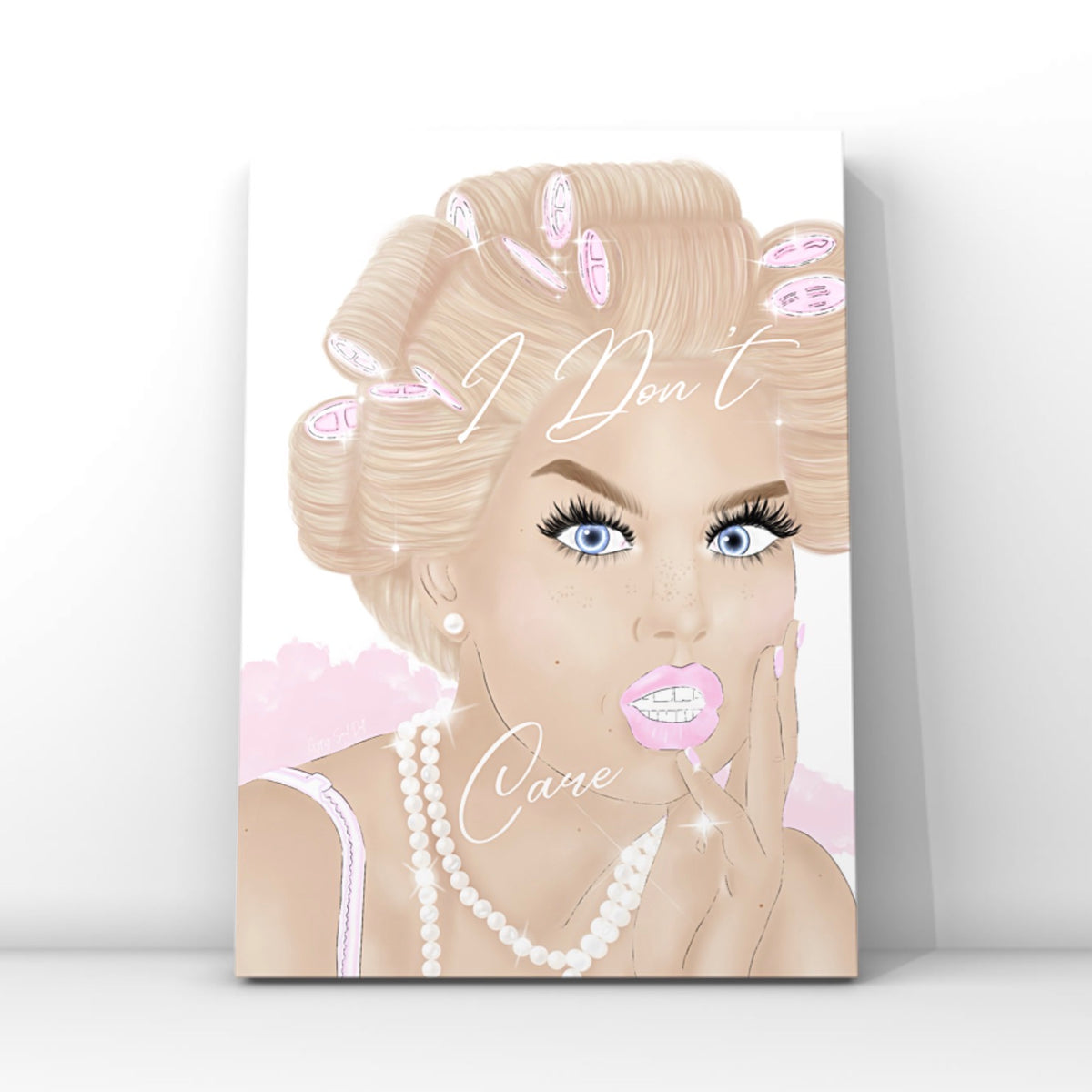 A vertical wall art print featuring a stylised illustration of a blonde woman with pink hair curlers, blue eyes, long lashes and pink lips, with the words 'I Don't Care' written across the portrait by Gypsy Soul Doll. The glam print is shown leaning against a white wall on a light grey floor.