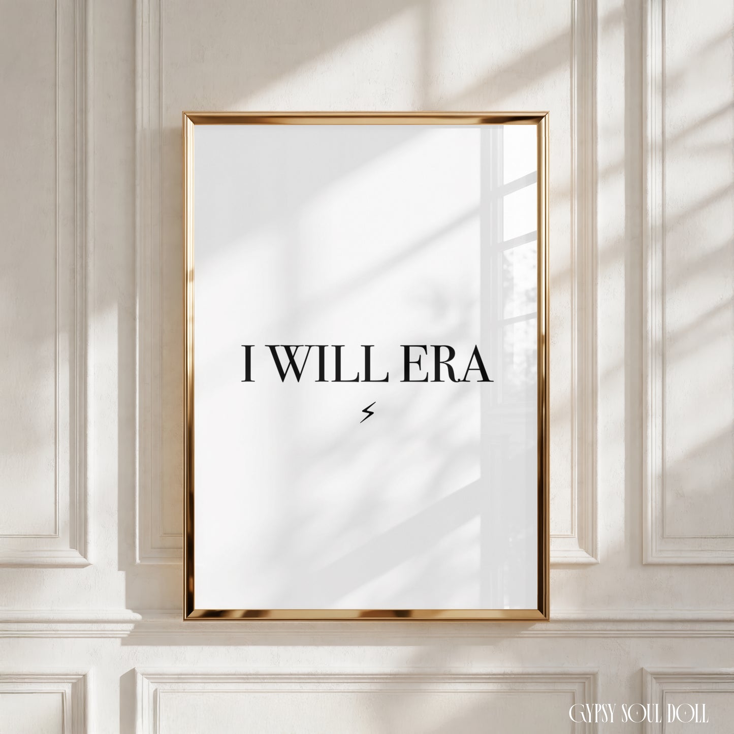 A framed art print with the text 'I WILL ERA' in black lettering on a white background, enclosed in a thin gold frame and leaning against a white panelled wall with soft window shadows.