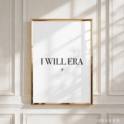 A framed art print with the text 'I WILL ERA' in black lettering on a white background, enclosed in a thin gold frame and leaning against a white panelled wall with soft window shadows.