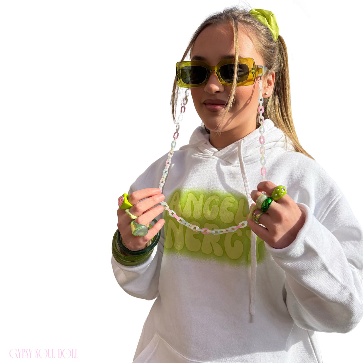A young woman with a ponytail and green sunglasses holds a beaded necklace with a green pendant in front of a white hoodie with 'ANGEL ENERGY' printed on the chest.