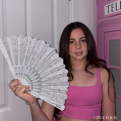 Model with long brown hair holding open the Gypsy Soul Doll Ibiza folding hand fan, showcasing the detailed white lace mesh material and shimmering silver glitter filigree scrollwork.