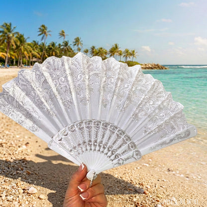 A white lace folding hand fan held by a hand against a tropical beach backdrop with turquoise water, sand, and palm trees.