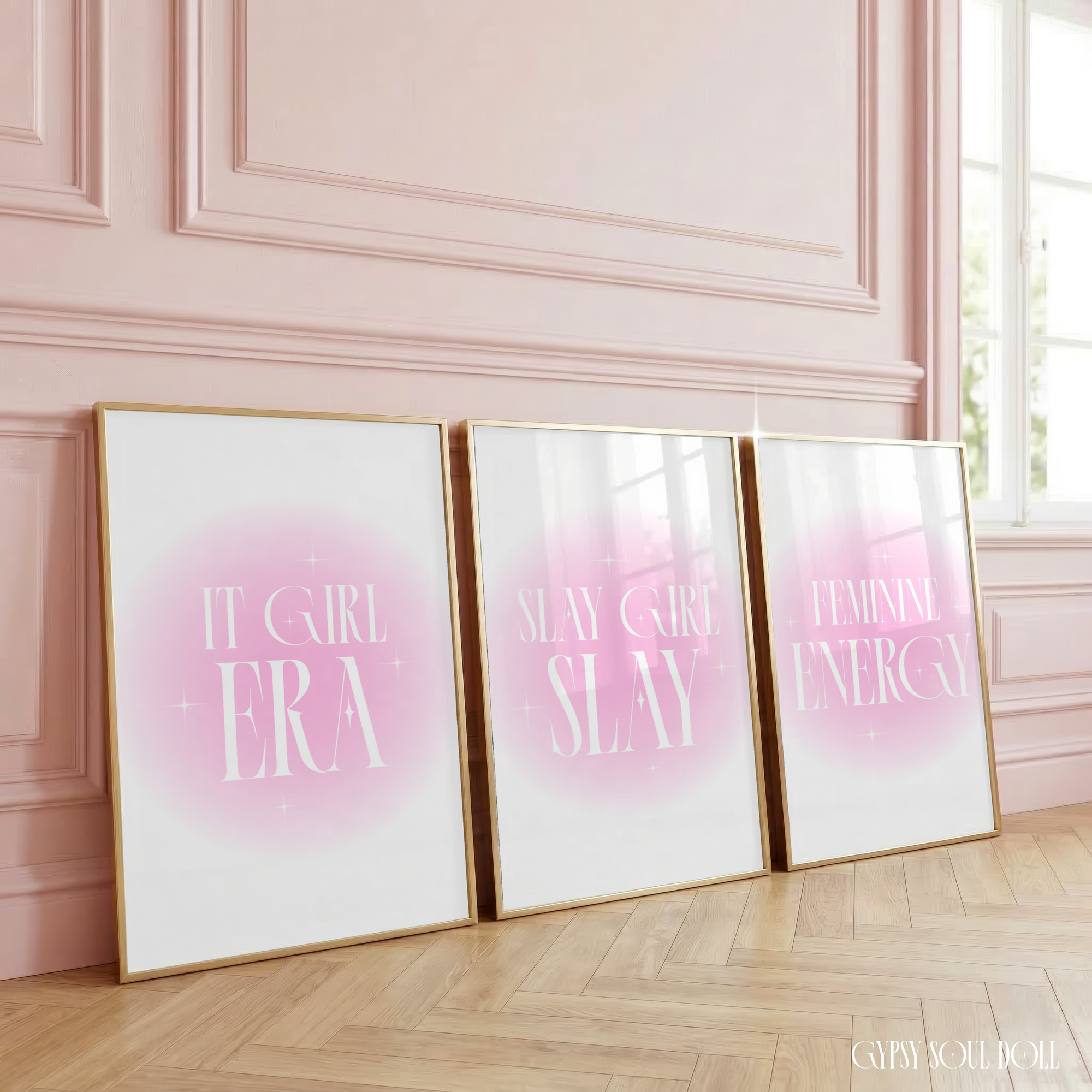Three matching minimalist typography prints, including It Girl Era, Slay Girl Slay, and Feminine Energy, with tall white serif fonts in glowing pink aura gradient backgrounds, styled in gold frames, leaning against a pink panelled wall.