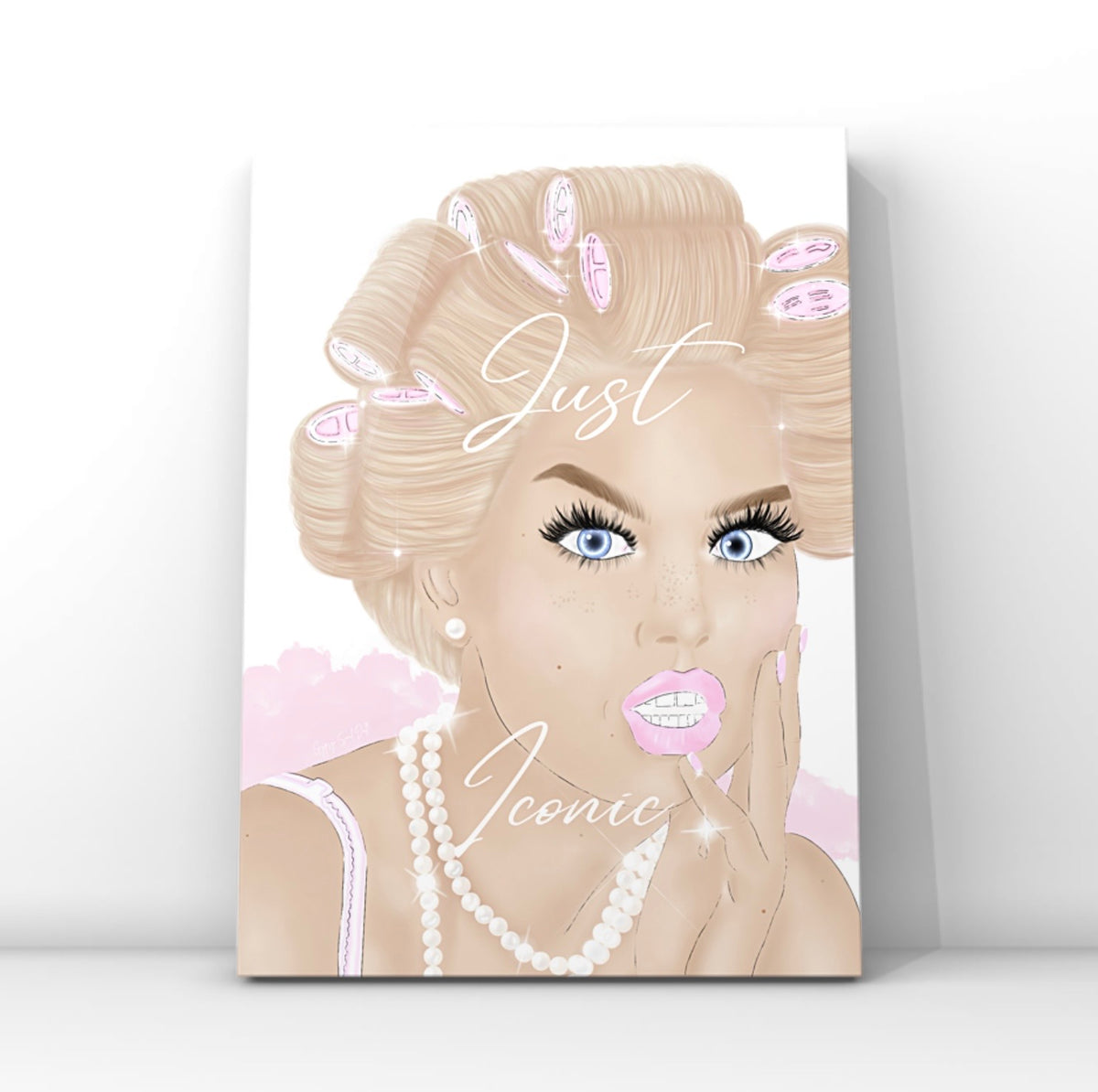 A vertical wall art print featuring a stylised illustration of a blonde glam woman with pink hair curlers, blue eyes, long lashes and pink lips, with the words 'Just Iconic' written across the centre, shown leaning against a white wall by Gypsy Soul Doll.