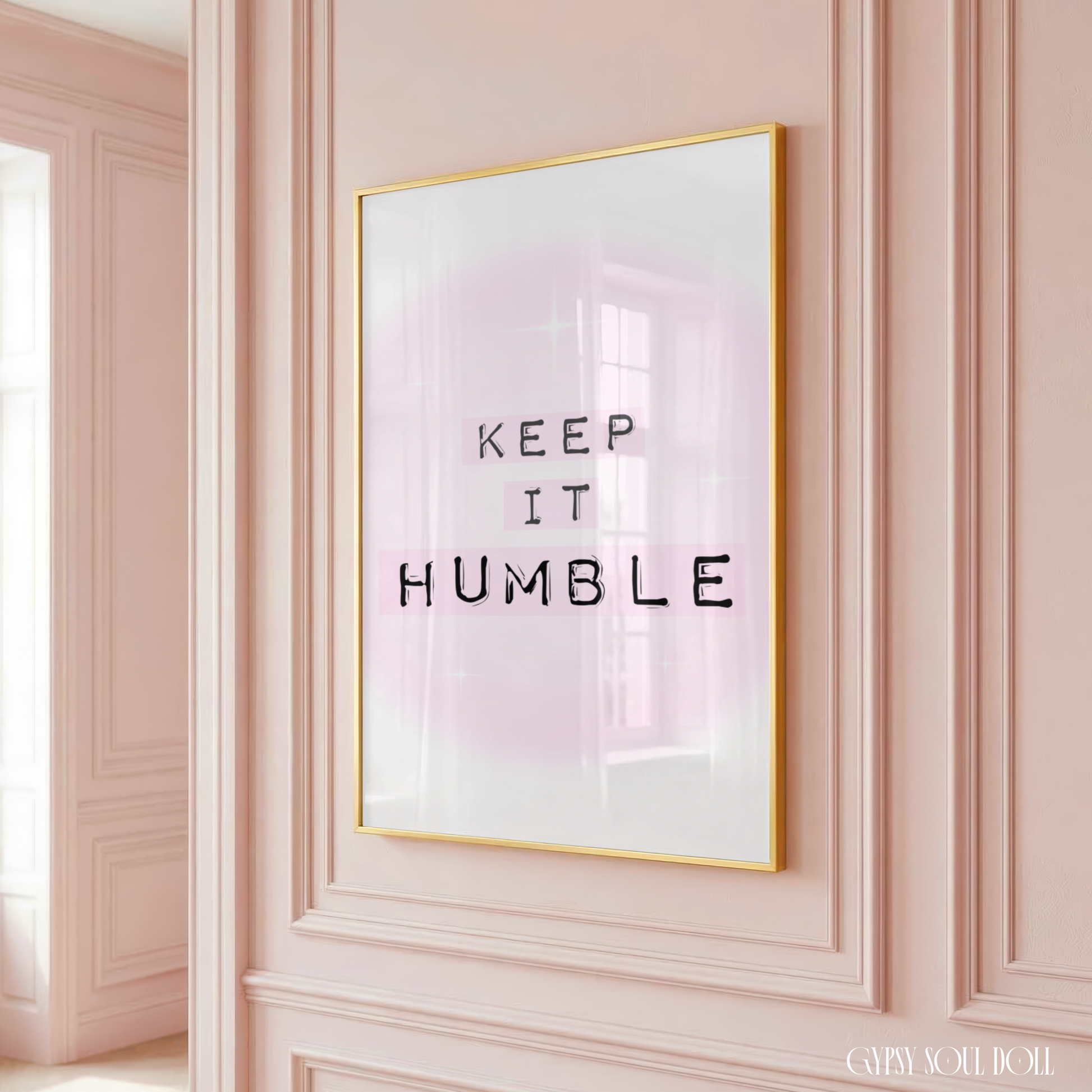 Minimalist A4 typographic wall art print featuring the phrase KEEP IT HUMBLE in bold black lettering highlighted by soft pastel pink blocks over a dreamy pink aura gradient background, framed on a wooden floor against a pink panelled wall!