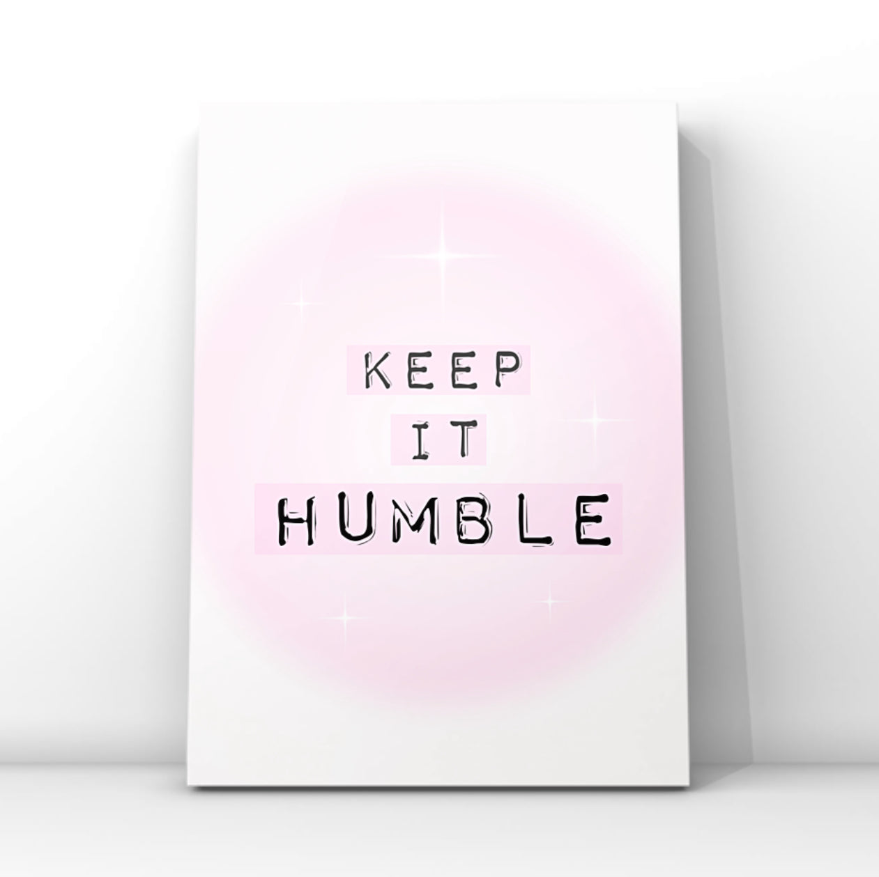 Minimalist A4 typographic wall art print featuring the phrase KEEP IT HUMBLE in bold black lettering highlighted by soft pastel pink blocks over a dreamy pink aura gradient background, framed on a wooden floor against a pink panelled wall!
