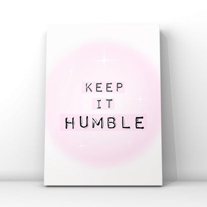Minimalist A4 typographic wall art print featuring the phrase KEEP IT HUMBLE in bold black lettering highlighted by soft pastel pink blocks over a dreamy pink aura gradient background, framed on a wooden floor against a pink panelled wall!