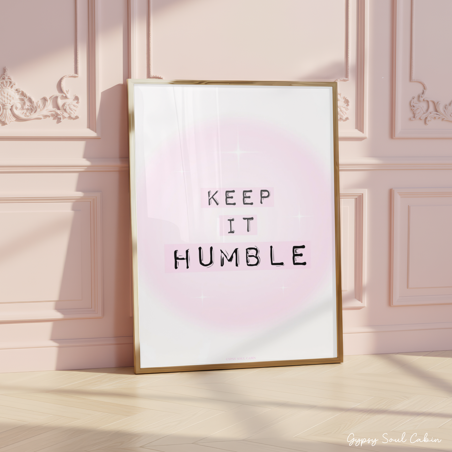Minimalist A4 typographic wall art print featuring the phrase KEEP IT HUMBLE in bold black lettering highlighted by soft pastel pink blocks over a dreamy pink aura gradient background, framed on a wooden floor against a pink panelled wall.