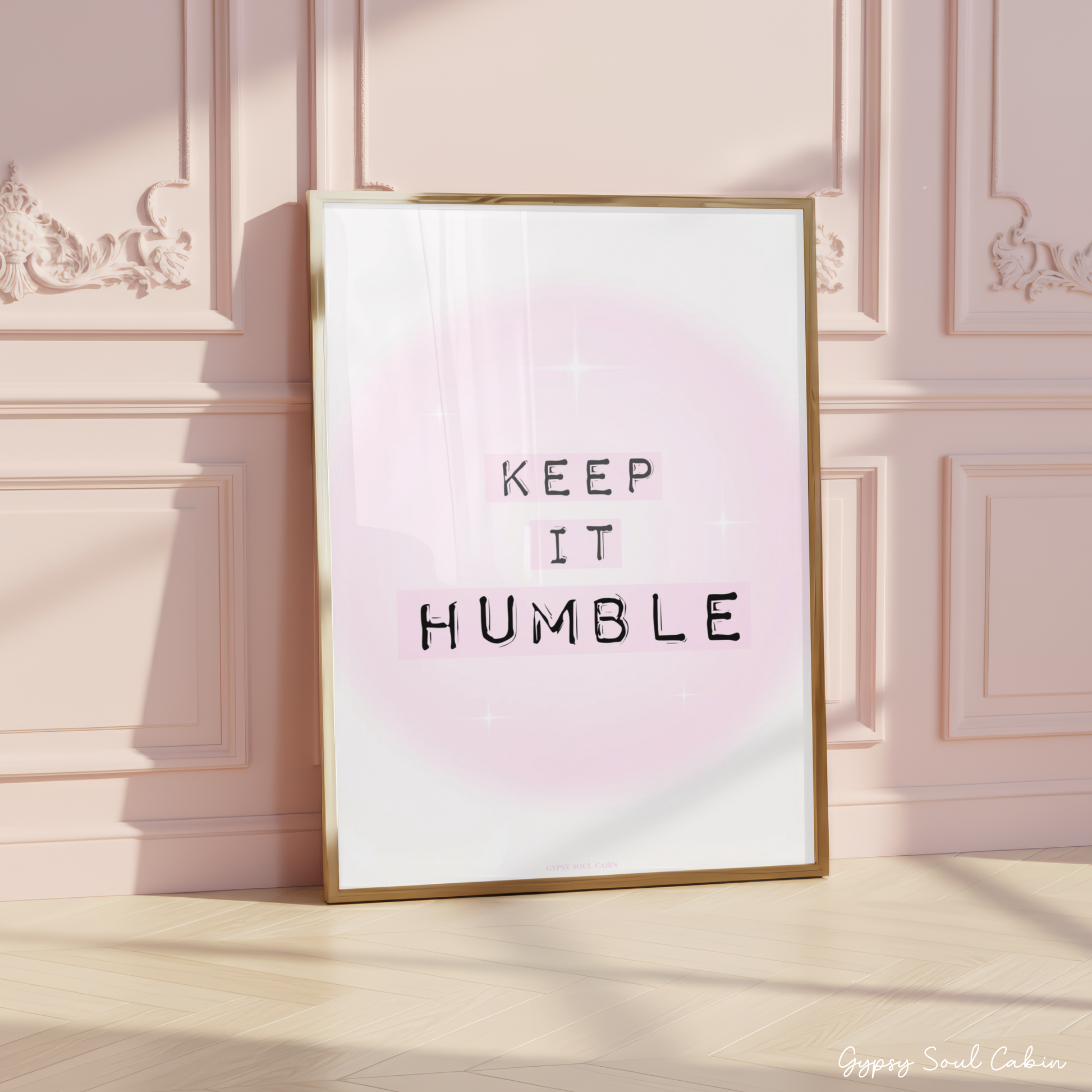 Minimalist A4 typographic wall art print featuring the phrase KEEP IT HUMBLE in bold black lettering highlighted by soft pastel pink blocks over a dreamy pink aura gradient background, framed on a wooden floor against a pink panelled wall.