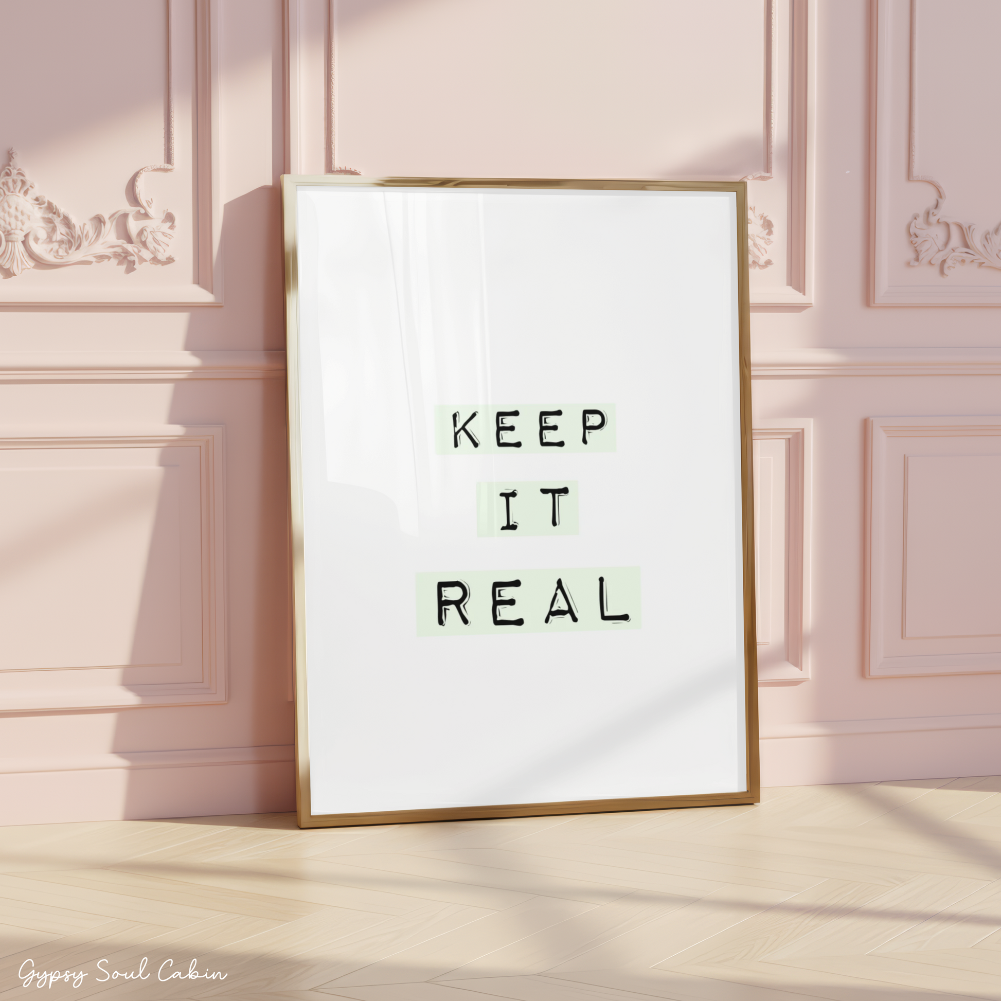 Minimalist A4 typographic wall art print featuring the phrase KEEP IT REAL in black typewriter font highlighted by soft pastel green blocks on a clean white background, framed on a wooden floor against a pink panelled wall.