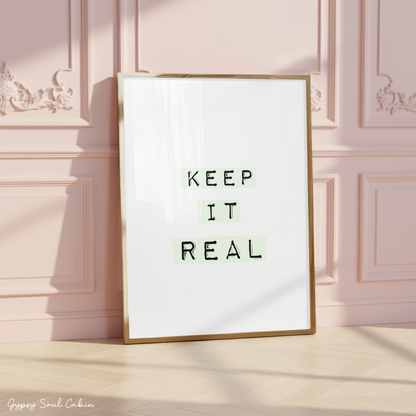 Minimalist A4 typographic wall art print featuring the phrase KEEP IT REAL in black typewriter font highlighted by soft pastel green blocks on a clean white background, framed on a wooden floor against a pink panelled wall.