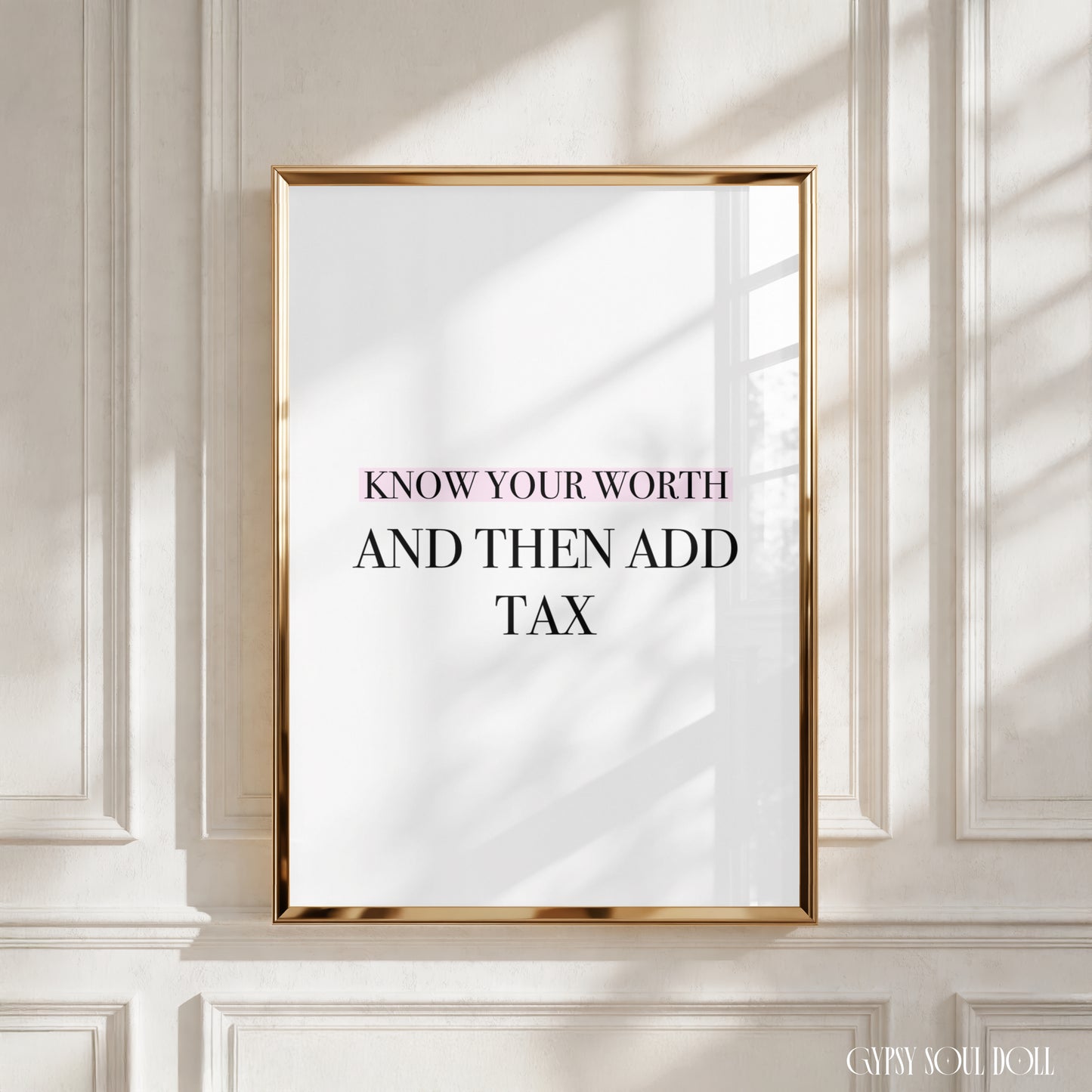 A framed art print with the text 'KNOW YOUR WORTH AND THEN ADD TAX' in black lettering on a white background, enclosed in a thin gold frame and leaning against a white paneled wall with soft window shadows.