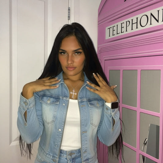 A woman posing in front of a pink telephone booth, wearing a light blue denim jacket with distressed details over a white top.
