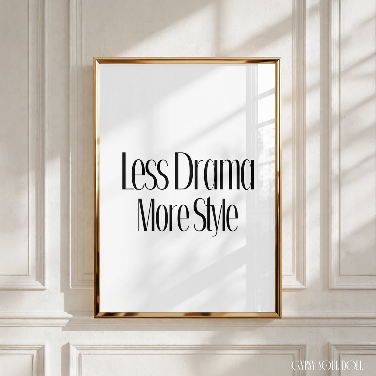A vertical framed wall art print with the text 'Less Drama More Style' in black serif lettering on a white background, set in a thin gold frame and mounted on a white panelled wall with soft window shadows.